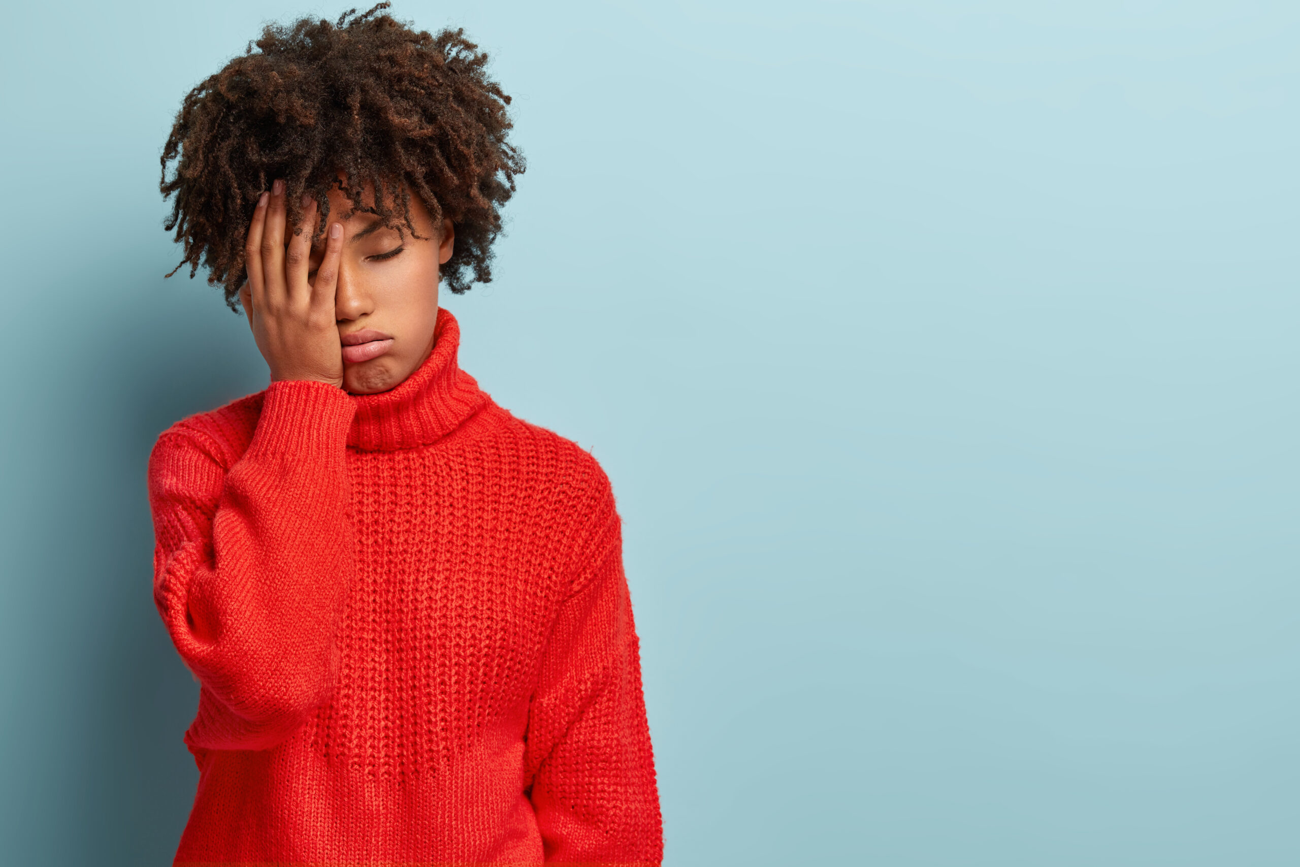 Discontent tired dark skinned woman covers face with hand, has tired expression, wears red sweater, isolated over blue wall with empty space for your promotion. Fatigue overworked female student
